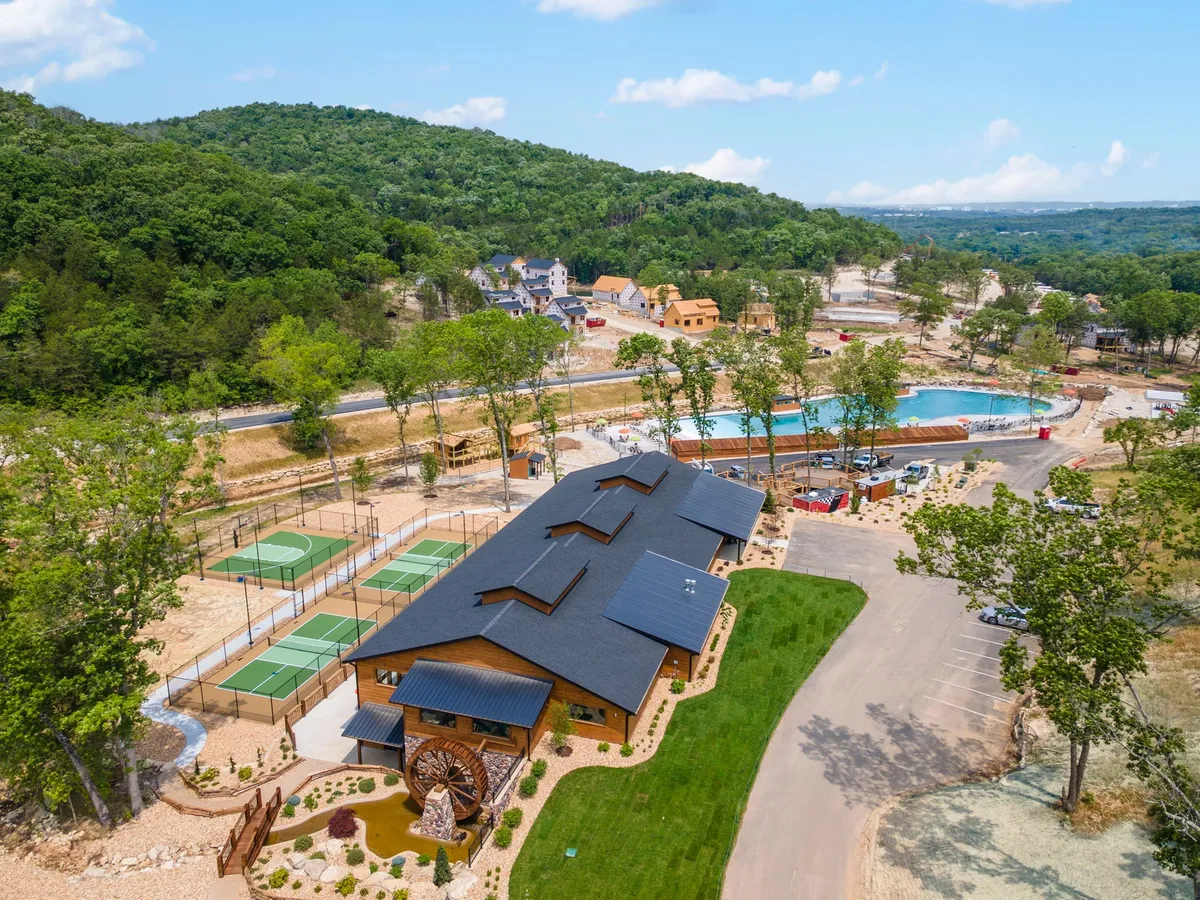 This exterior image features the main building of the resort, a large swimming pool, tennis courts, and the surrounding natural landscape. The scene is vibrant and bustling, suggesting recreational opportunities and community events. The pool area is inviting, with lounge chairs set up beside the water, and the tennis courts signal additional active pursuits for families.