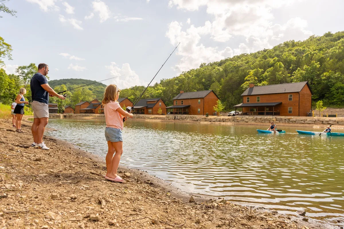 A family enjoys fishing by the sparkling lake, with kayakers paddling in the background and rustic wooden cabins nestled among rolling green hills. The lakeside scene conveys a sense of adventure, tranquility, and the opportunity for memorable shared moments in nature.