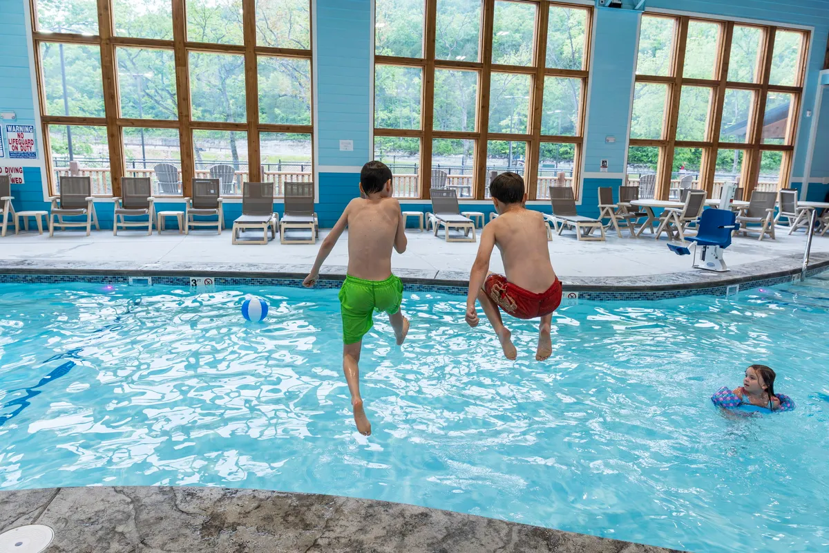 This vibrant image captures two boys joyfully jumping into a sunlit indoor swimming pool, while a girl swims nearby. Large windows flood the space with natural light, highlighting the playful energy and family-friendly ambiance. Poolside lounge chairs and scattered beach balls enhance the carefree, resort-like atmosphere.