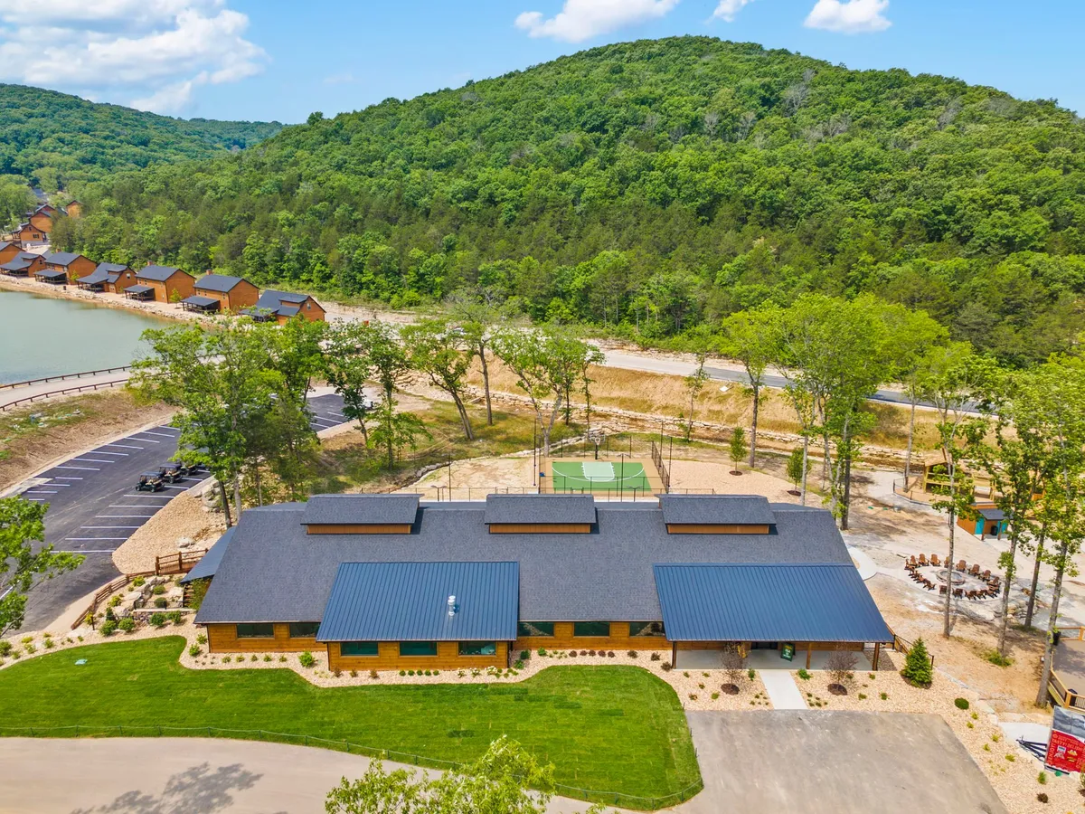 The scene showcases the main building, lush natural surroundings, an outdoor sports area, and cozy cabins set by the water. The setting is relaxed and communal, with opportunities for fishing, sports, and nature walks along the lakeside.