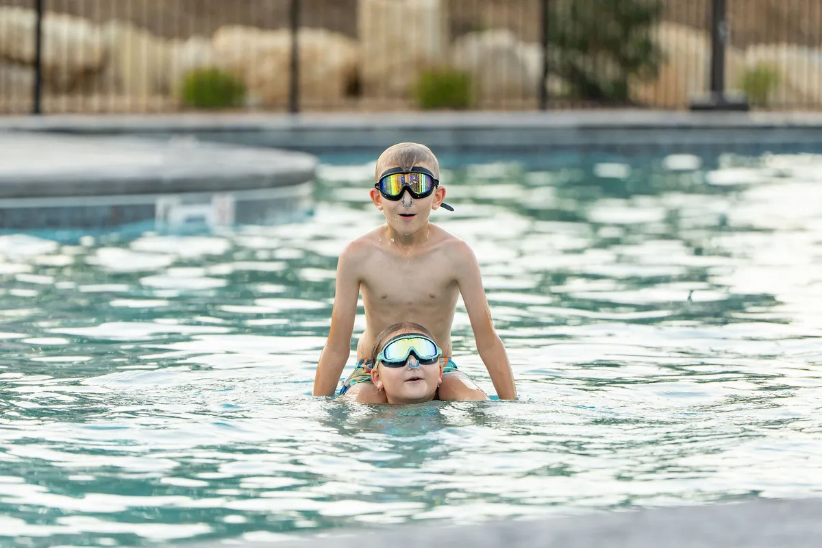 Two children are joyfully playing in an outdoor swimming pool, both wearing goggles and splashing water. The reflections in the pool and the bright natural light evoke a sense of carefree summer days and family bonding.