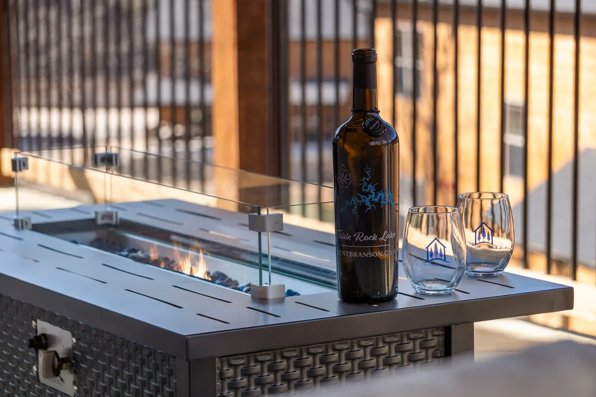 An inviting fire pit area with a bottle of wine and glasses arranged on a rustic table, surrounded by Adirondack chairs and trees, capturing the essence of outdoor gatherings at sunset.