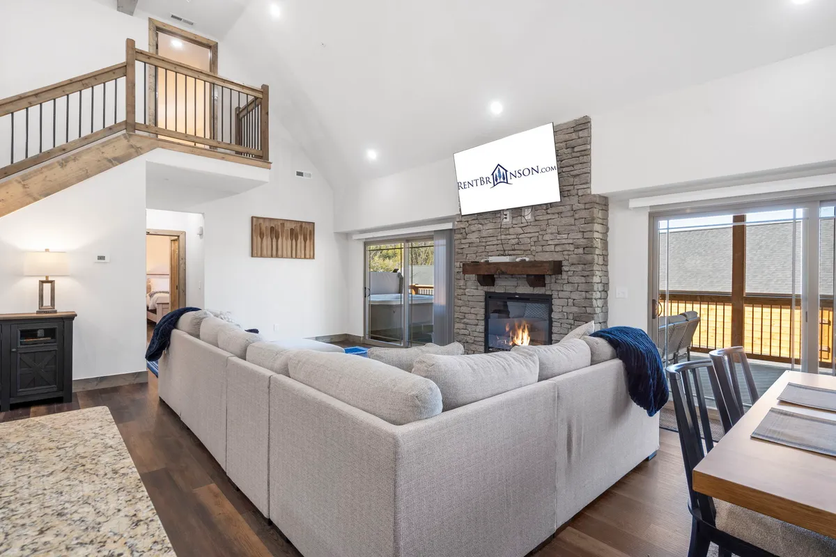 This image showcases a spacious open-concept living area with a large sectional sofa, a cozy fireplace, a dining table, a staircase, and expansive windows bringing in natural light. The room has a warm, inviting atmosphere ideal for group gatherings and relaxation after a day of adventure.