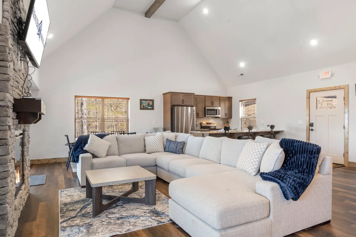 A spacious living room with a large stone fireplace, an inviting sectional sofa, and a view into the adjacent modern kitchen area. The scene radiates comfort and togetherness, making it ideal for family game nights, meals, or simply unwinding.